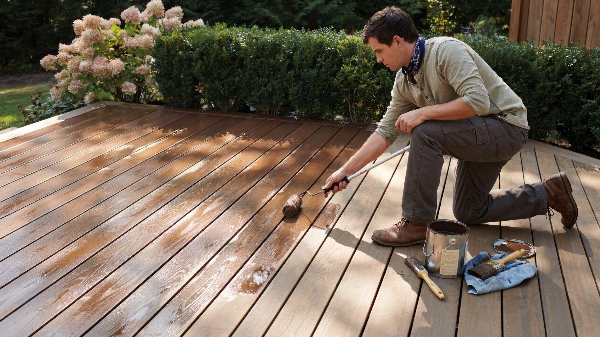 Freshly stained wooden deck drying in sunlight with tools nearby as a homeowner applies a second coat in one section for better coverage.