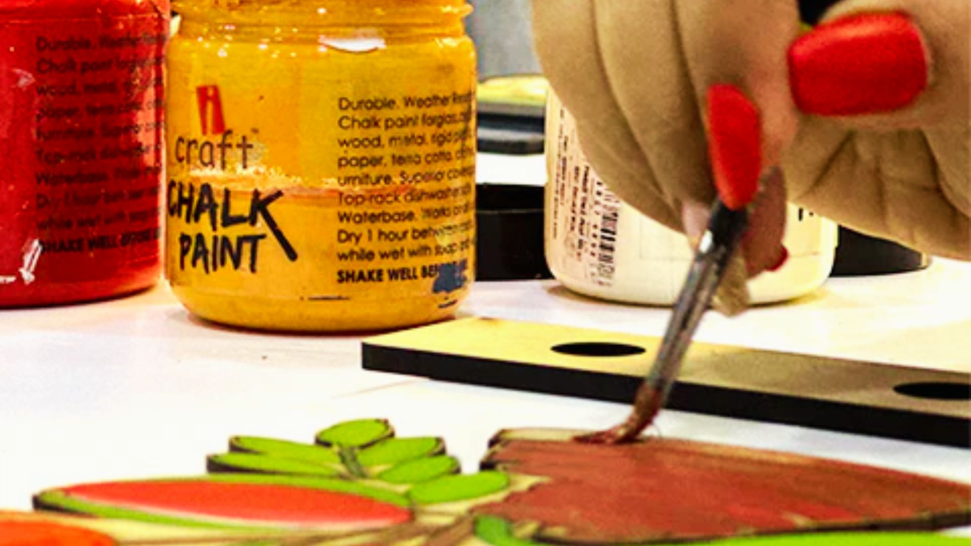 Woman using a small brush to apply chalk paint on a wooden craft piece next to iCraft chalk paint jars.