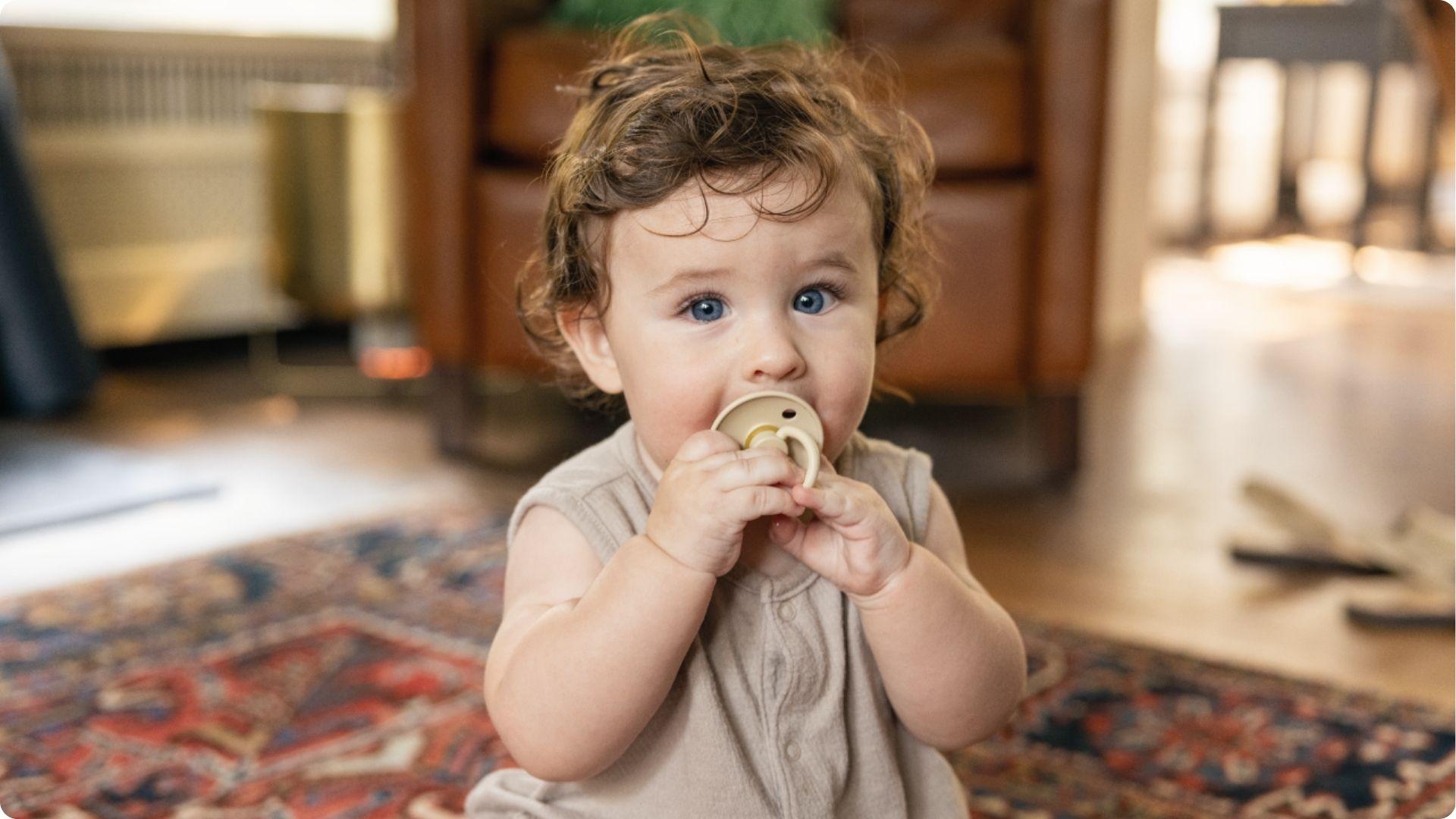 Blue-eyed toddler holds a beige pacifier, showing common night time soother habit before weaning starts.