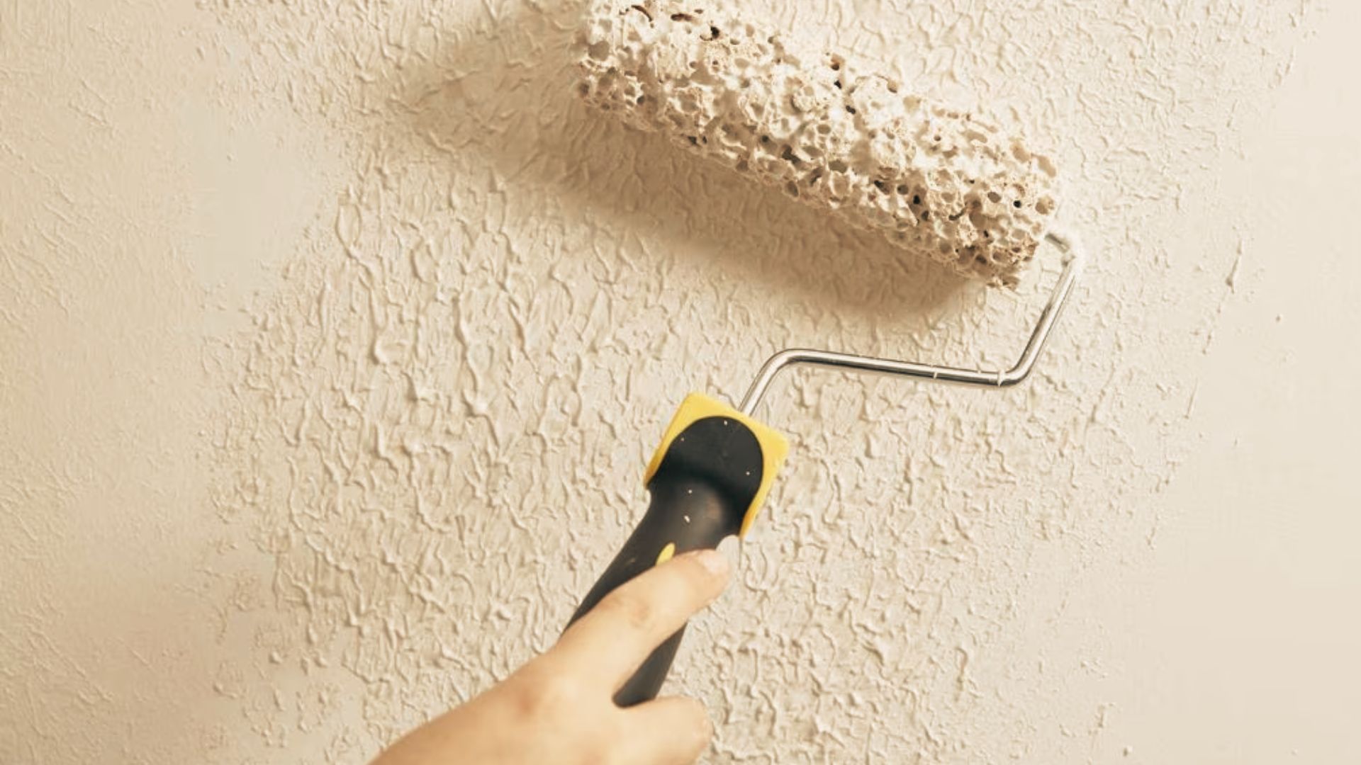 Person using a paint roller to apply thick textured cream paint onto a wall with an uneven finish.