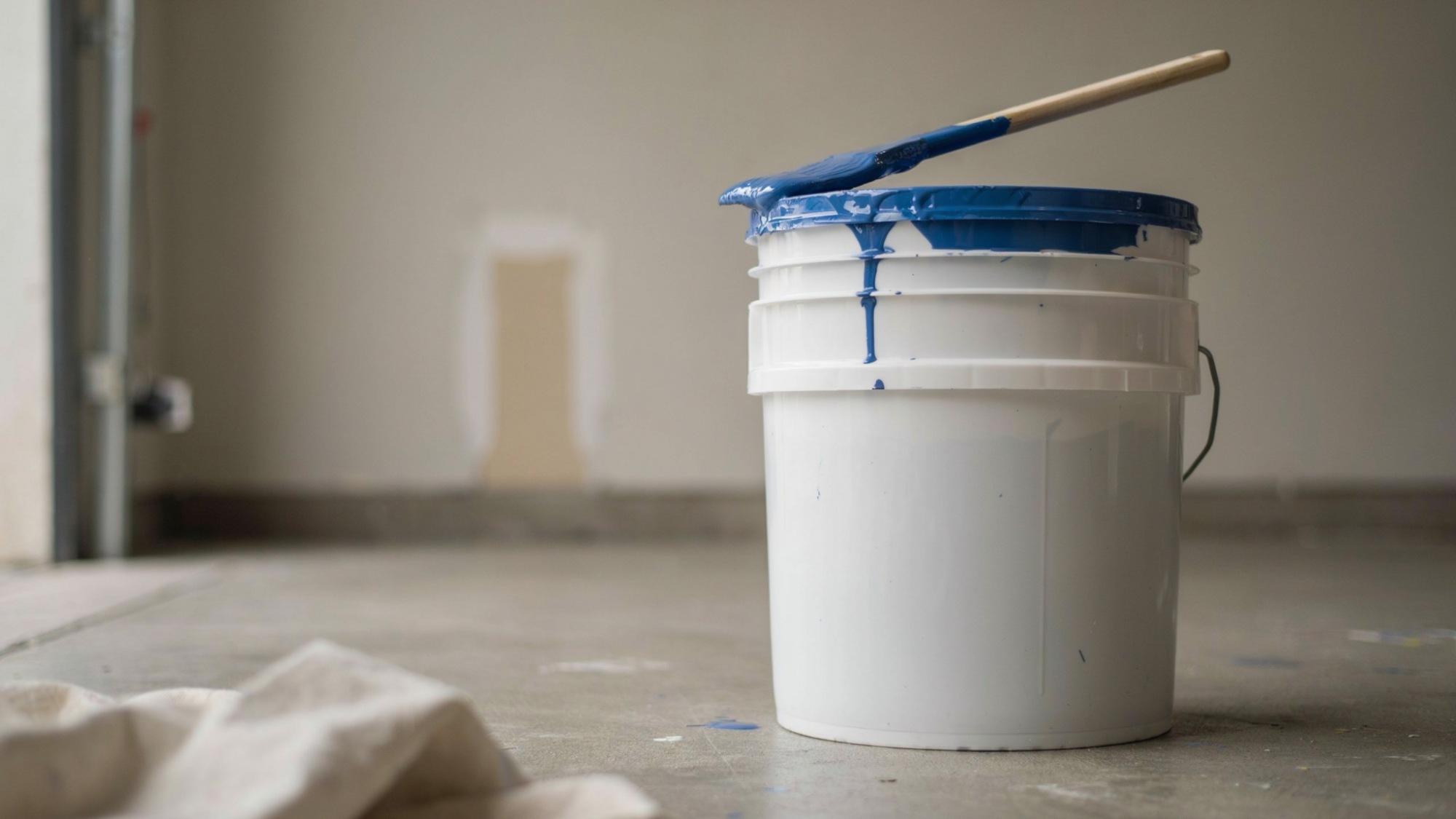 White paint bucket with blue paint dripping from lid and wooden mixing stick on floor.