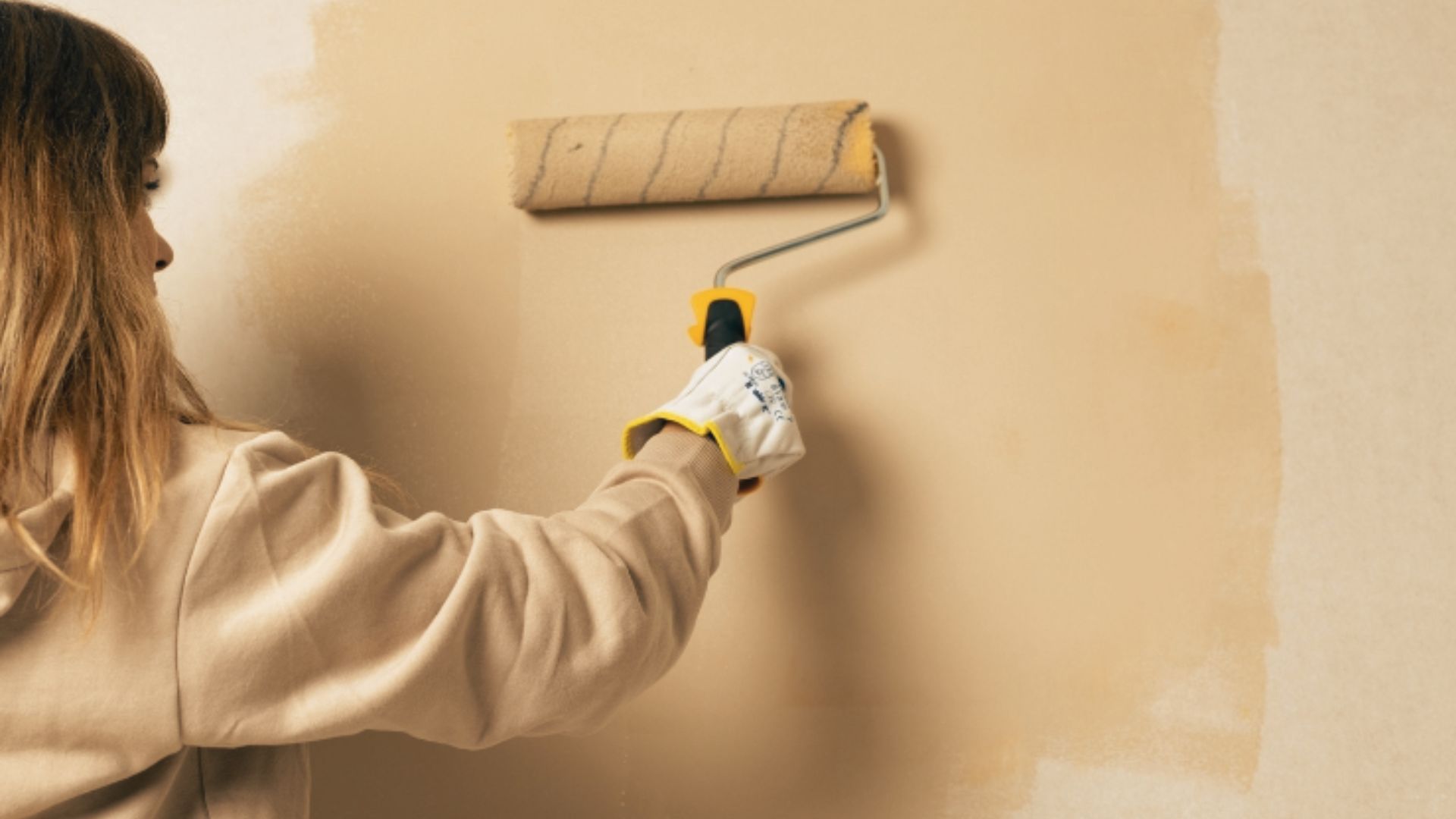 Woman rolling beige paint onto a wall wearing protective gloves and a cream coloured sweater indoors.