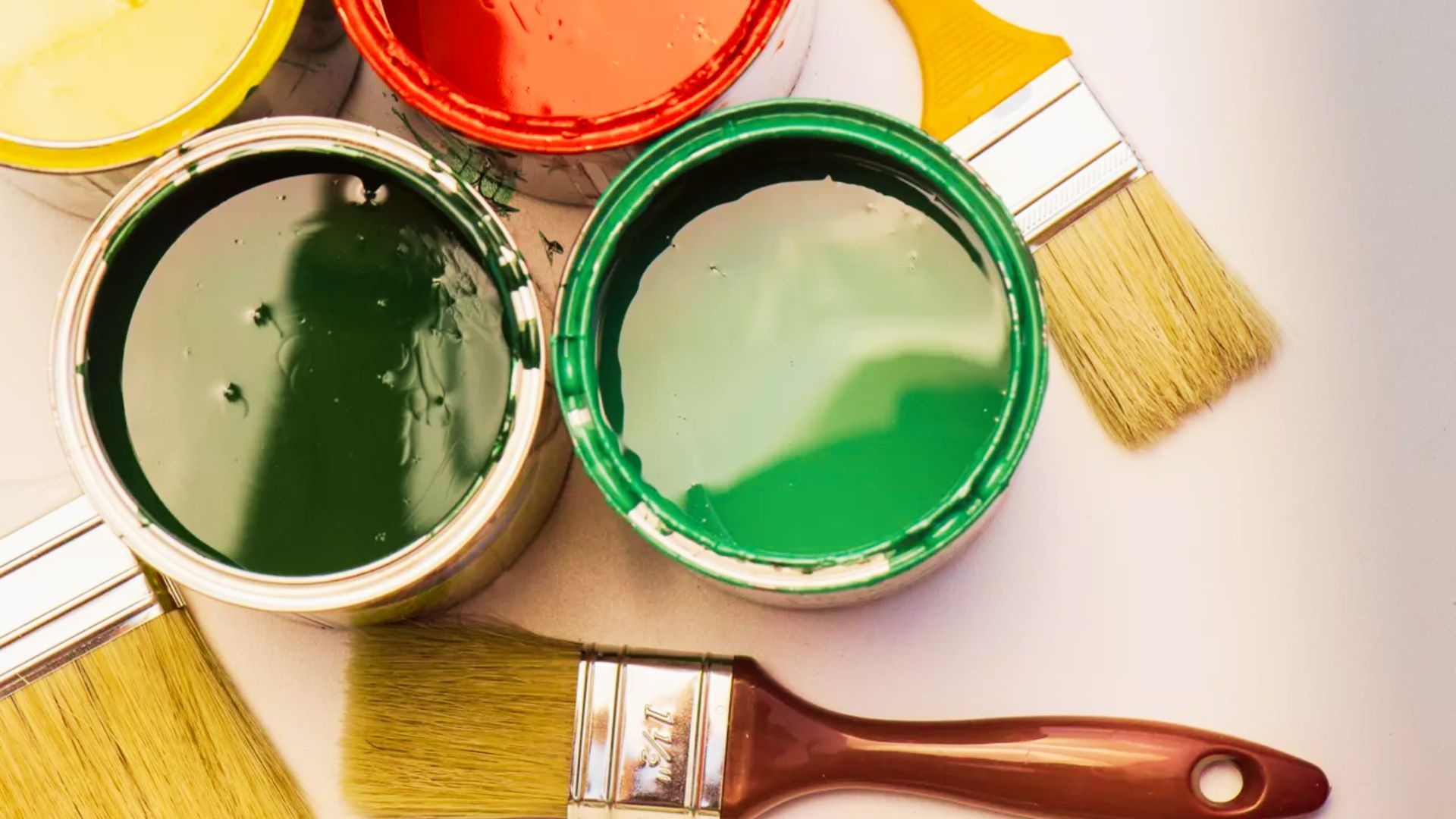 Top-down view of open paint cans in green, red, and yellow with wooden-handled brushes on a white surface.