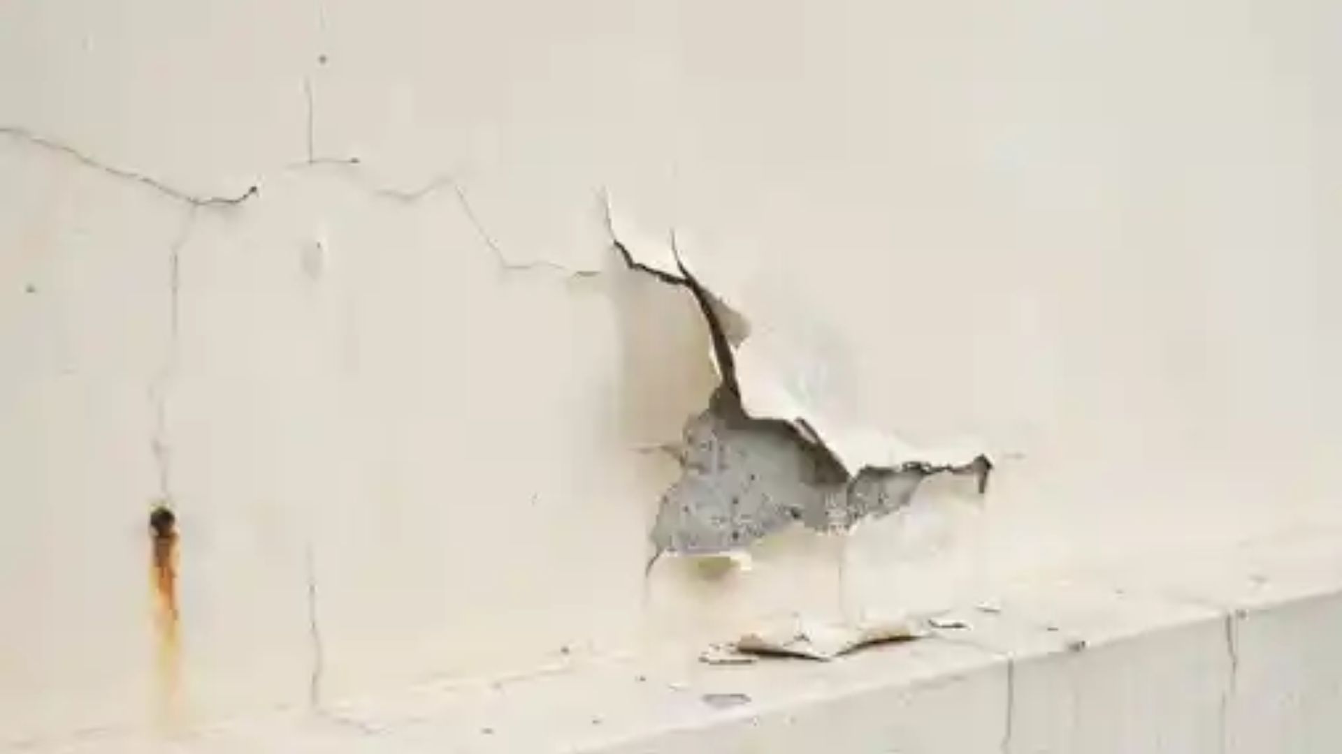 Damaged white wall with peeling and cracking paint showing rust stains and exposed grey surface underneath.