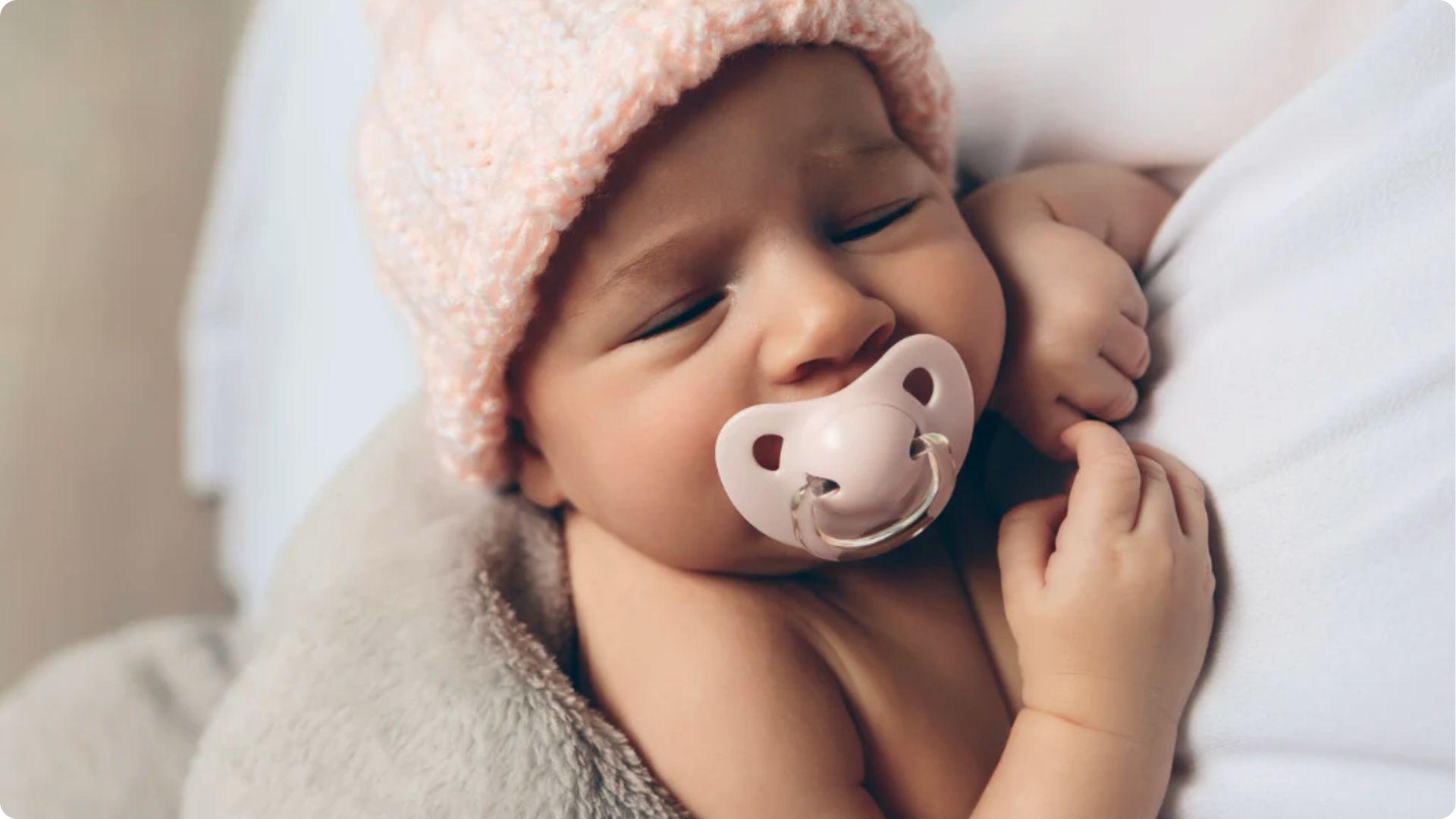 Newborn sleeps with a pink pacifier and knit hat, representing an early habit to address for night weaning.