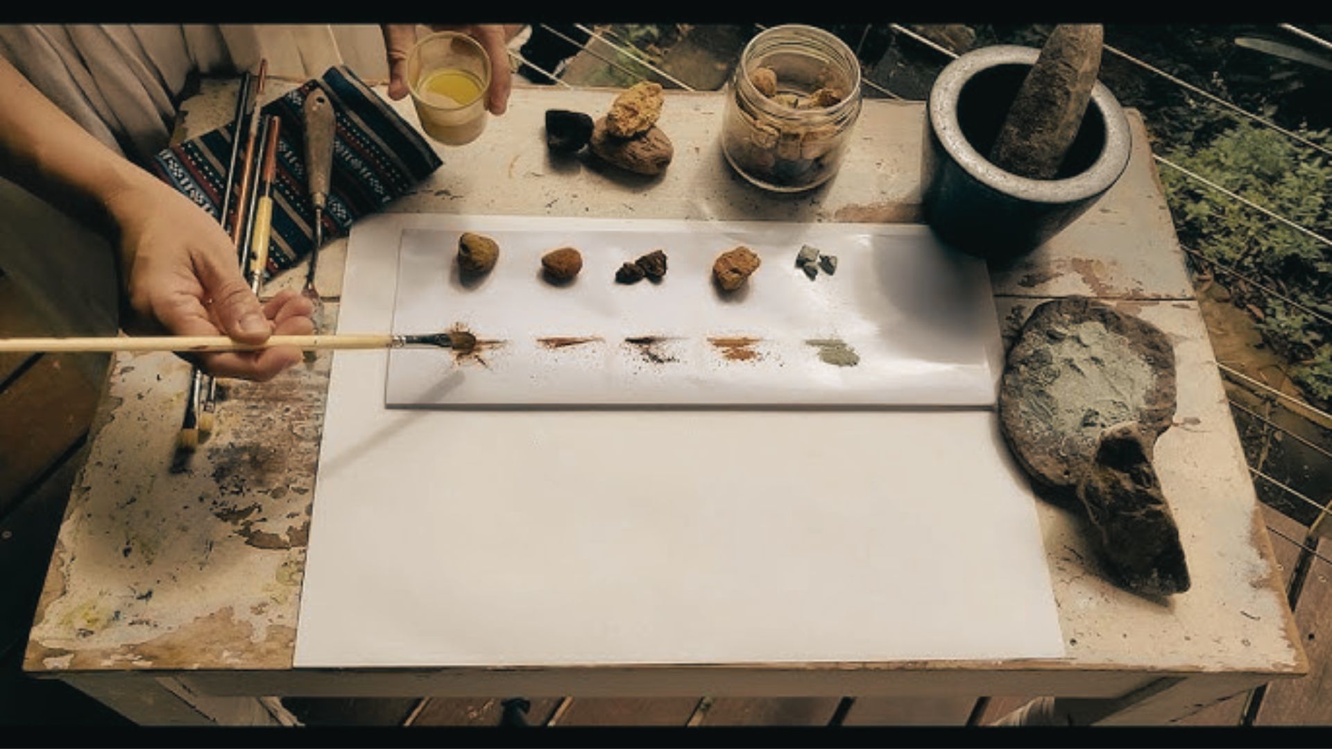 Hands grinding natural pigments from rocks on paper, with brushes and mortar nearby.