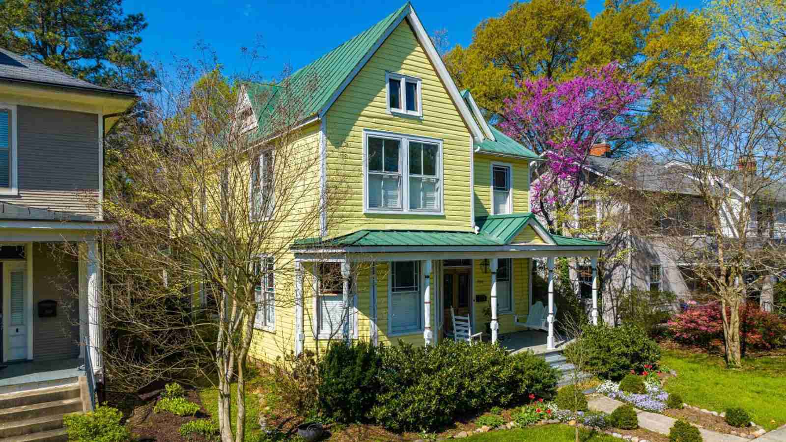 Two-story pale yellow Victorian house with green metal roof, white porch columns, rocking chairs, landscaped garden and blooming purple tree.